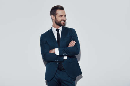 Handsome businessman. Charming young man keeping arms crossed and smiling while standing against grey backgroundの写真素材