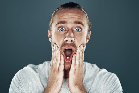 Unbelievable! Shocked young man looking at camera and making a face while standing against grey...