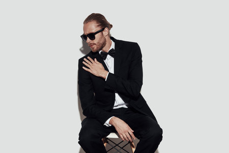 Checking details. Charming young man in full suit cleaning jacket while sitting against grey...
