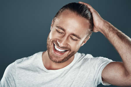 Cheerful. Good looking young man keeping hand in hair and smiling while standing against grey backgroundの写真素材