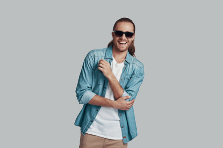Young and carefree. Handsome young man looking at camera and smiling while standing against grey...