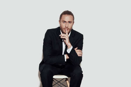 Keep my secret! Handsome young man in full suit looking at camera and keeping finger on lips while sitting against grey backgroundの写真素材