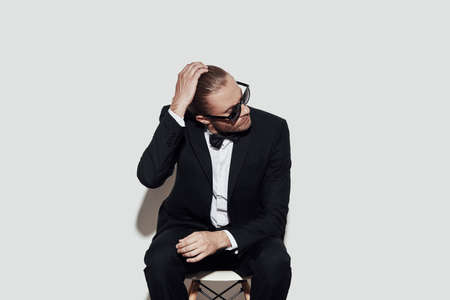 Positively charming. Charming young man in full suit adjusting hair while sitting against grey backgroundの写真素材