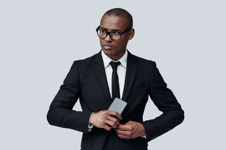 Young and smart. Charming young African man in formalwear using smart phone while standing against grey backgroundの写真素材