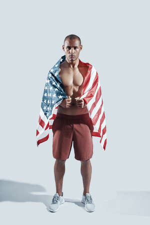 Strength and power. Full length of handsome young African man looking at camera and covering with American flag while standing against grey backgroundの写真素材