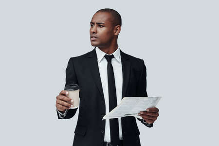Aware of latest news. Handsome young African man in formalwear holding disposable cup and newspaper while standing against grey backgroundの写真素材