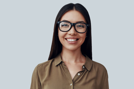 Intelligence. Attractive young Asian woman looking at camera and smiling while standing against grey...