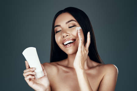 Feeling great. Beautiful young Asian woman smiling and applying moisturizer while standing against grey backgroundの写真素材