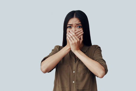 Oh no! Scared young Asian woman covering mouth with hands while standing against grey background