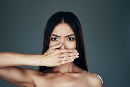 Secret. Beautiful young Asian woman looking at camera and covering mouth with hand while standing against grey backgroundの写真素材