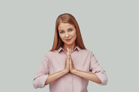 Attractive young woman keeping hands clasped and looking at camera while standing against grey...