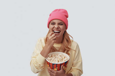 Attractive young woman eating popcorn and smiling while standing against grey backgroundの写真素材