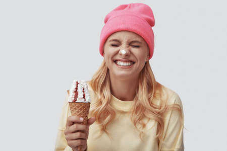 Attractive young woman eating ice cream and smiling while standing against grey backgroundの写真素材
