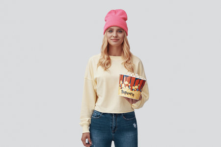 Attractive young woman holding popcorn and smiling while standing against grey background