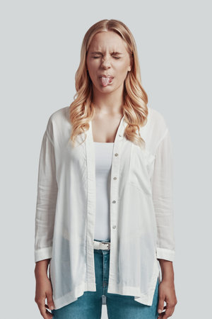Attractive young woman making a face and sticking out tongue while standing against grey background