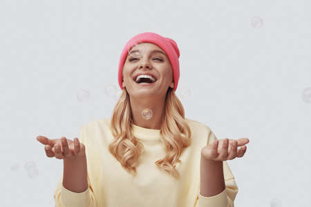 Attractive young woman playing with soap bubbles and smiling while standing against grey backgroundの写真素材