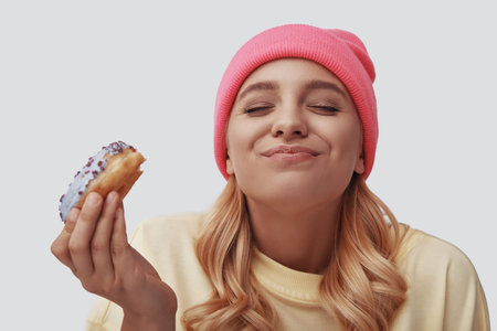 Attractive young woman eating donut and smiling while standing against grey background