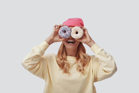 Attractive young woman covering eyes with donuts and smiling while standing against grey background