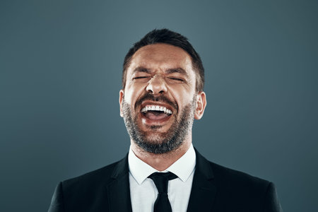 Charming young man in full suit laughing while standing against grey background