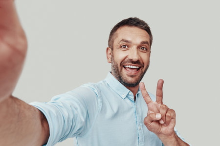 Self portrait of handsome young man looking at camera and smiling while standing against grey...