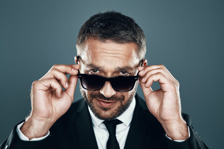 Charming young man in full suit adjusting eyewear while standing against grey background