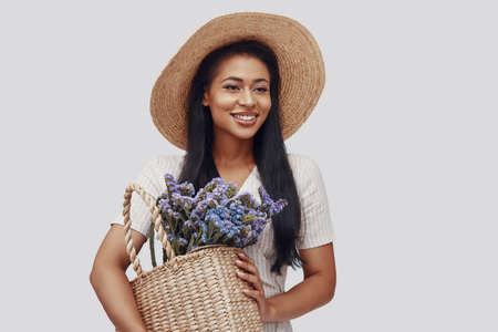 Attractive young woman in hat smiling and carrying basket with flowers while standing against grey backgroundの写真素材