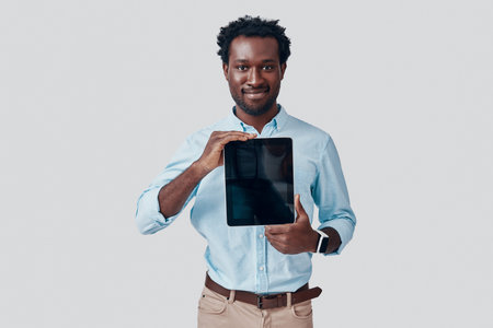 Handsome young African man pointing copy space on digital tablet and smiling while standing against grey backgroundの写真素材