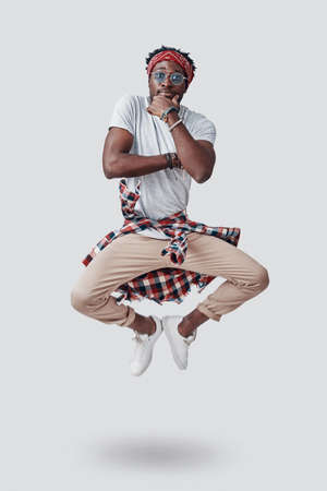 Full length of playful young African man looking at camera and keeping hand on chin while hovering against grey backgroundの写真素材