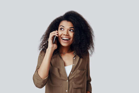 Beautiful young African woman talking on the smart phone and smiling while standing against grey backgroundの写真素材