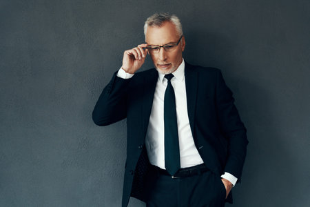 Handsome senior man in full suit adjusting eyewear while standing against grey background