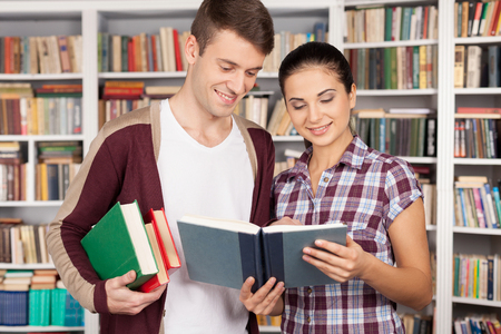 Studying together is fun. Cheerful young woman and man standing close to each other and reading book at the libraryの写真素材