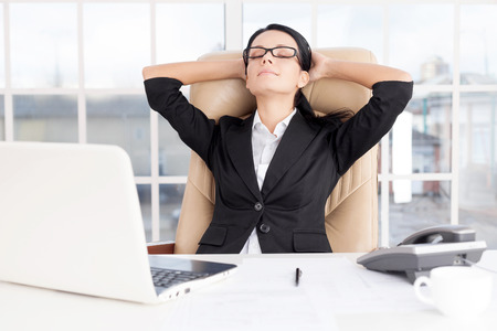 Chill time. Cheerful young business woman holding head in hands and keeping eyes closed while sitting at her working placeの写真素材