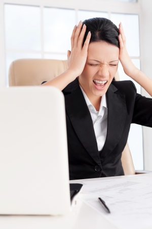 Emotional stress. Young business woman holding head in hands and shouting while sitting at her working placeの写真素材