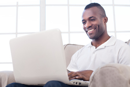 Working at home. Cheerful African man using computer and smiling while sitting on the chairの写真素材