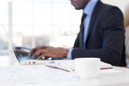 Businessman at work. Close-up of African businessman using computer with a coffee cup on the foregroundの写真素材