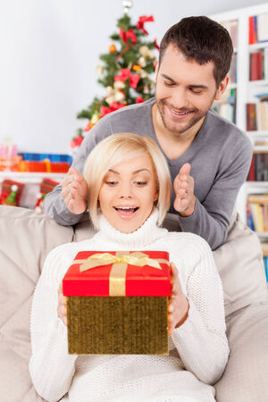 Surprise! Beautiful young woman sitting on the couch and holding a gift box while her boyfriend standing behind her and smilingの写真素材