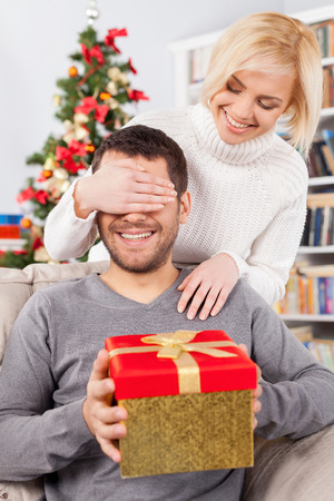 Surprise! Handsome young man sitting on the couch and holding a gift box while her girlfriend standing behind him and covering his eyes with handsの写真素材