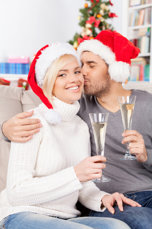 Christmas kiss. Beautiful young woman holding champagne flute and smiling while her boyfriend kissing her cheekの写真素材