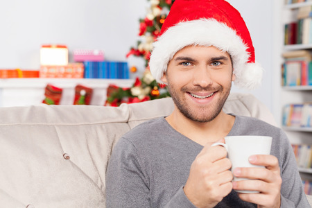 Hot drink on Christmas Eve. Cheerful young man in Christmas hat holding a cup and smiling at cameraの写真素材