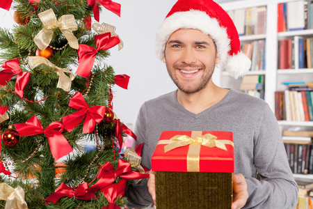 Present for you. Handsome young man holding a gift box and smiling at camera while standing near the Christmas treeの写真素材