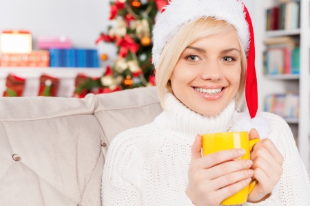 Hot drink on Christmas Eve. Beautiful young woman in Christmas hat holding a cup and smiling at cameraの写真素材