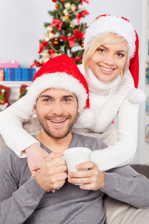 Together on Christmas Eve. Cheerful young man in Christmas hat holding a cup and smiling while his girlfriend standing behind and hugging himの写真素材