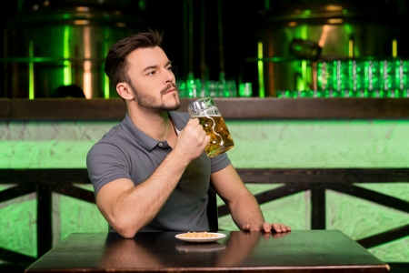 Drinking fresh beer. Thoughtful young man holding a mug with beer and looking away while sitting in barの写真素材