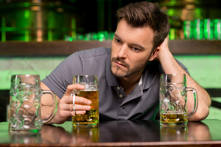 Lonely man in bar. Depressed young man drinking beer and holding hand in hair while sitting in bar の写真素材