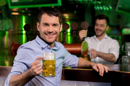 Man in beer pub. Handsome young man holding a beer mug and smiling while bartender pouring beer on the backgroundの写真素材