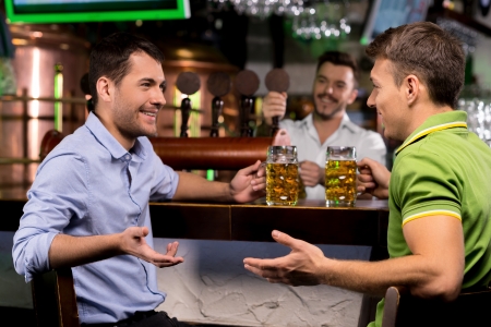 Spending time in bar. Two young man drinking beer and talking to each other while bartender tapping beer on the backgroundの写真素材