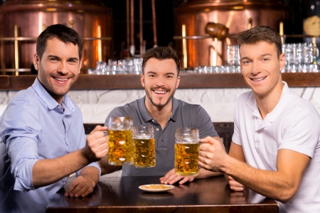 Having a good time. Three cheerful friends holding mugs with lager and smiling at cameraの写真素材