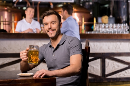 Man in beer pub. Cheerful young man holding a beer mug and smiling while sitting in barの写真素材