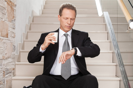 Late again. Worried mature man in formalwear holding a coffee cup and checking a time while sitting on staircase の写真素材