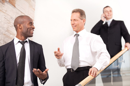 Business people on staircase. Three cheerful man in formalwear moving downstairs while two of them talking to each other and smilingの写真素材